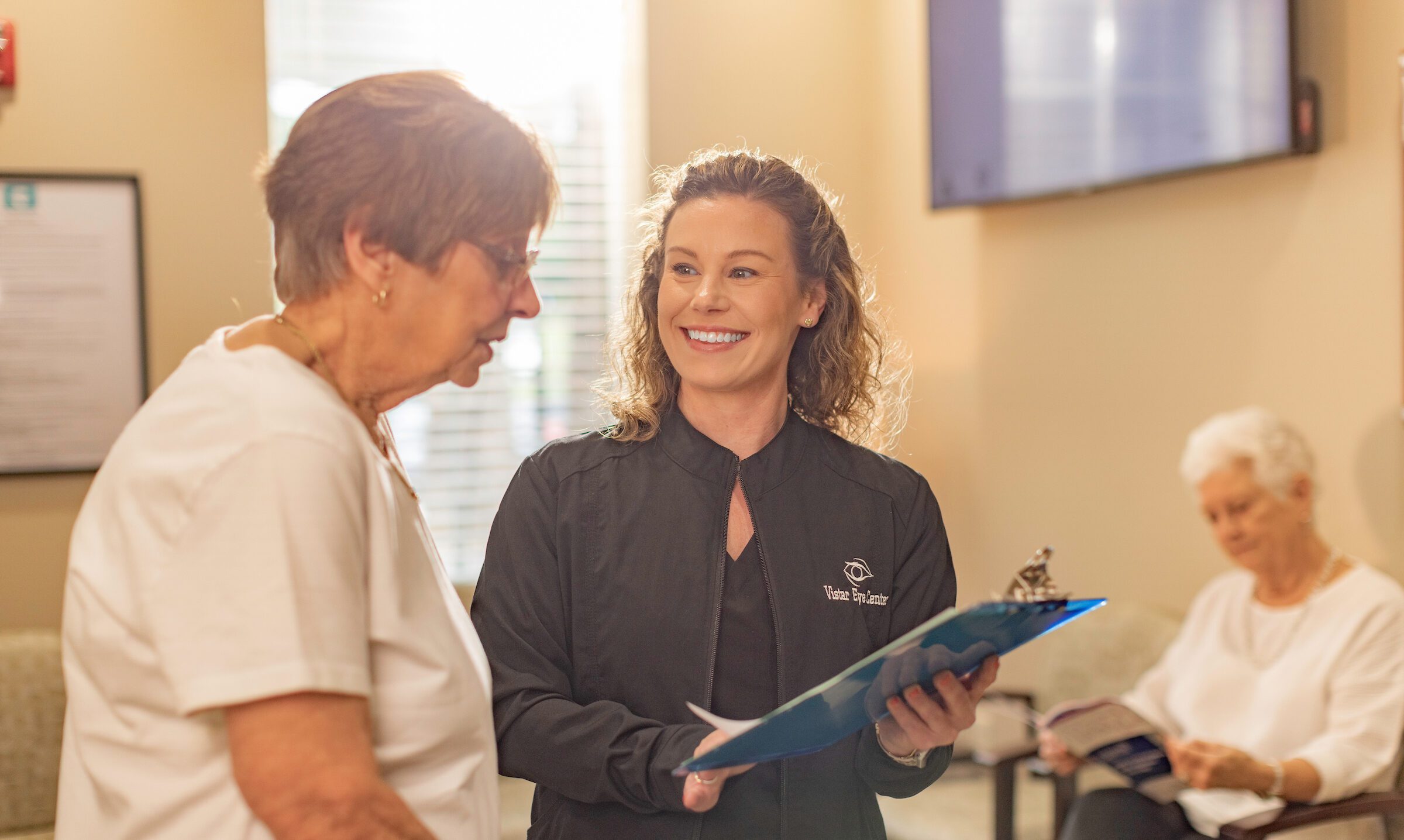 Staff member in black scrubs and Vistar zip-up holds a blue clipboard and smiles at an older female patient in the waiting room