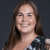 Headshot of a woman with long brown hair and a floral top smiling in front of a gray background